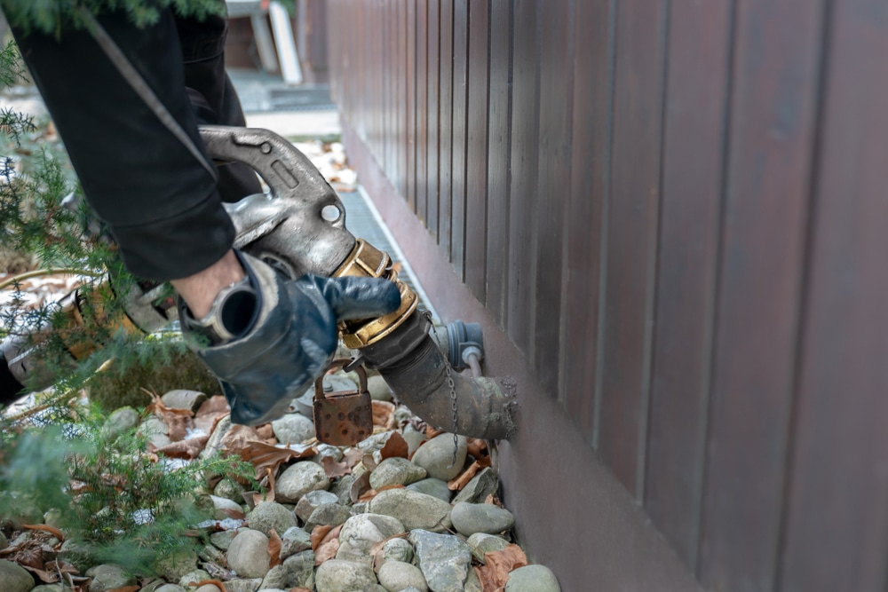 A worker in protective gloves connects a hose to an exterior heating oil tank fill pipe beside a building.